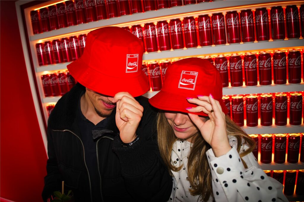 Deux invités essayant des bobs rouges Coca-Cola devant un mur de canettes