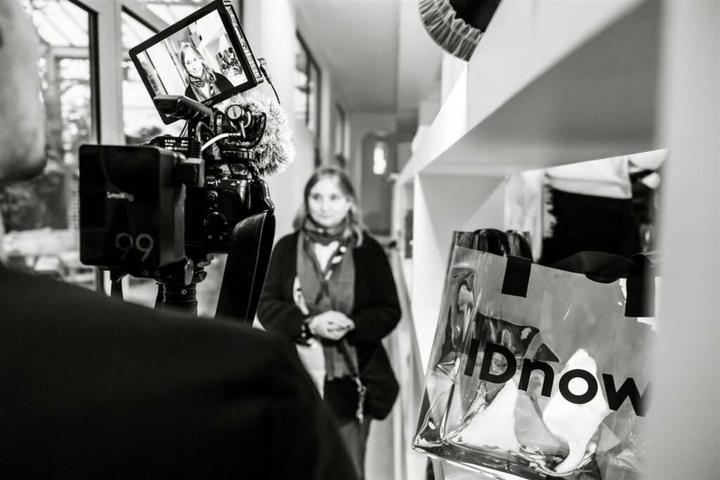 Coulisses en noir et blanc : caméra de tournage et invitée au second plan avec un sac IDnow